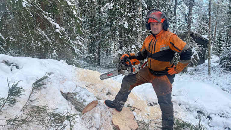 En man med skyddskläder står i en snötäckt skog med en motorsåg.