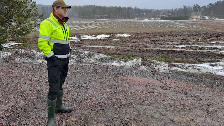 Man i gulsvarta kläder, keps och gummistövlar står vid en åkerkant.