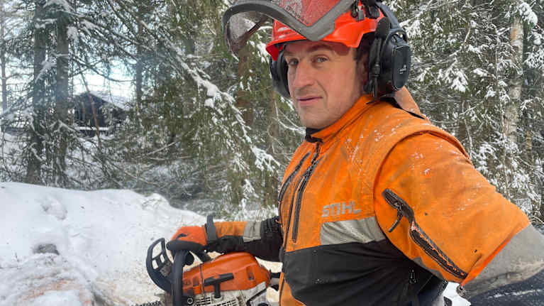 En man med skyddskläder står i en snötäckt skog med en motorsåg.
