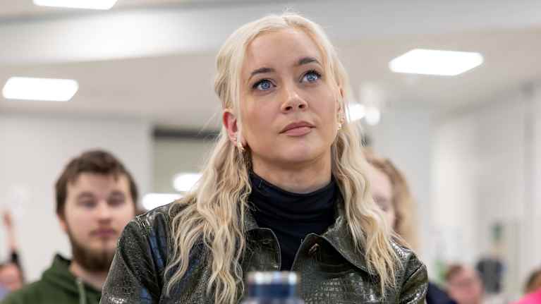 A blonde woman dressed in black sits at a desk looking forward.
