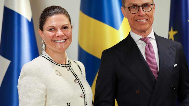Prinsessa Victoria valkoisessa jakussa, jossa hopeat napit, rinnassa ritakoru ohuesta kultalangasta ja helmistä. Vieressä seisoo Alexander Stubb.