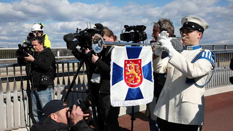 A man on a bridge, dressed in a white uniform playing a long horn instrument draped with a Finnish coat of arms flag, with media photographers in the background.