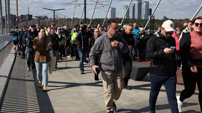 A large crowd of people walking on a bridge, with buildings, bridge cables and a few industrial smokestacks in the background.