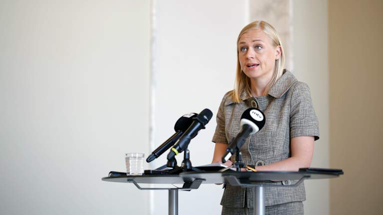 A blonde woman in a gray short-sleeved suit stood behind a microphone at a podium.