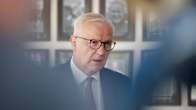 Olli Rehn puhuu. Hänellä on silmälasit, sininen puku, valkoinen kauluspaita ja punapilkullinen kravatti. Taustalla on tauluja.