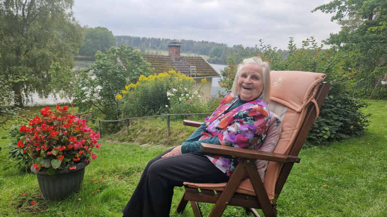 Vanha nainen istuu puutarhatuolissa puutarhassa ja nauraa kameralle.