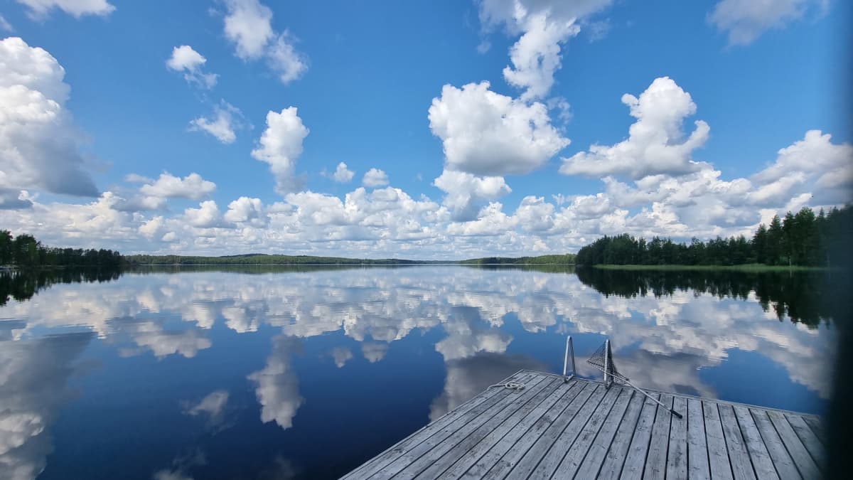 Järvimaisemaa. Tyyni päivä ja kumpupilvet heijastuvat järven pintaan. Laituri kurkottaa kohti järveä. 