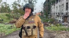 Jevgenij Prigozjin tar sig om hakan iklädd brunspräcklig skyddsväst och hjälm. Bakom honom syns en krigsskadad byggnad.