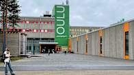 A young woman stands in front of a large grey concrete building with a large vertical green sign saying OULU University.