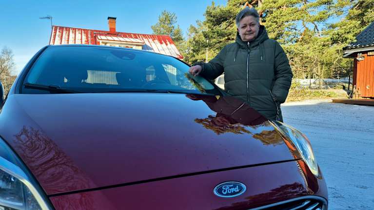 Äldre dam står bredvid sin rödmetallic Ford bil.