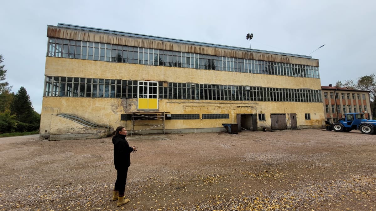 Keltainen iso suorakaiteenmallinen tehdasrakennus, jossa koko matkalta ikkunoita kahdessa kerroksessa. Rakennuksen edessä seisoo nainen mustissa vaatteissa. Oikealla kuvassa sininen kauhakuormaaja.  
