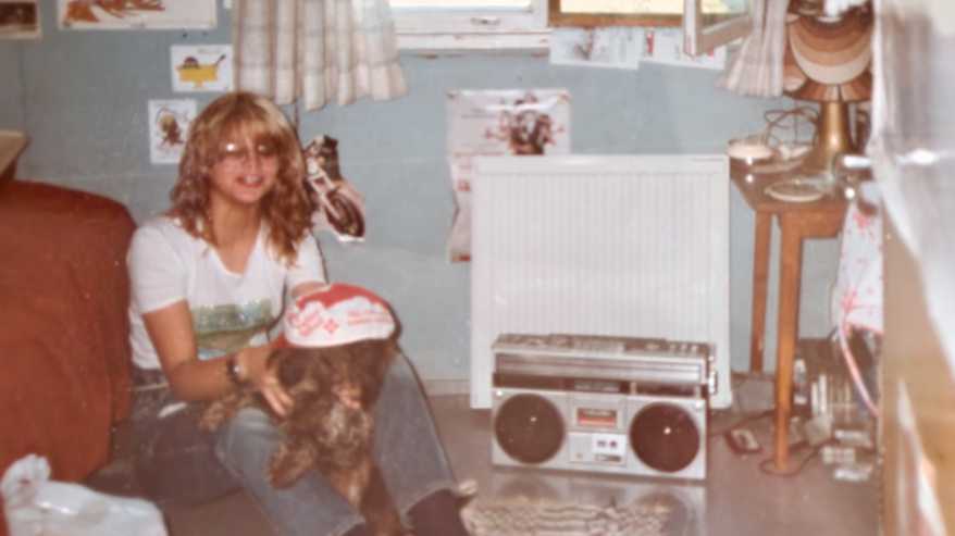 Vaalea silmälasipäinen nuori nainen, jolla on koira sylissä. Taustalla näkyy 80-luvun suuri stereomankka.