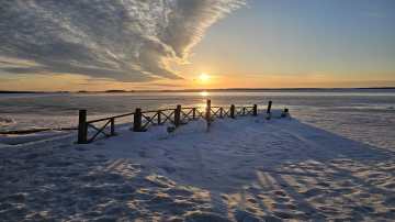 Aurinko paistaa horisontissa jäätyneen järven takaa. Laituri lumen ja jään keskellä. 