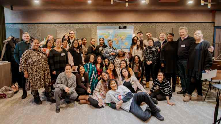 Iso joukko erilaisia ihmisiä vierekkäin poseeraamassa. Osa istuu lattialla. Taustalla kartta.