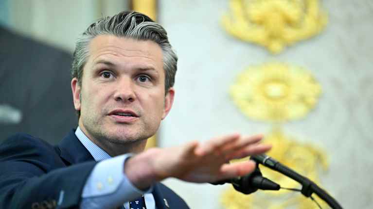 Pete Hegseth puhuu mikrofoniin oikea käsi ojennettuna eteenpäin. Hänellä on tummansininen puku ja kravatti ja vaaleansininen paita.
