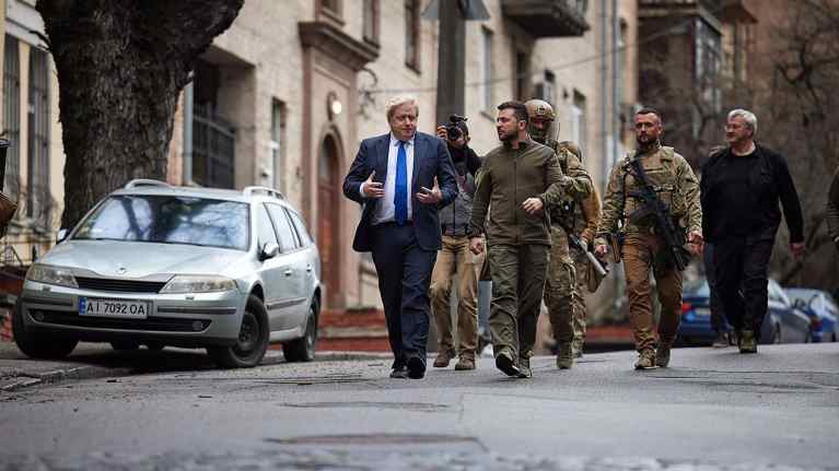 Boris Johnson ja Volodymyr Zelenskyi kävelevät kadulla. Johnsonilla on tumma puku ja sininen kravatti, Zelenskyillä khakinvihreä pusakka ja housut. Mukana kulkee valokuvaaja, ainakin kaksi aseistautunutta sotilasta ja musta-asuinen mies.