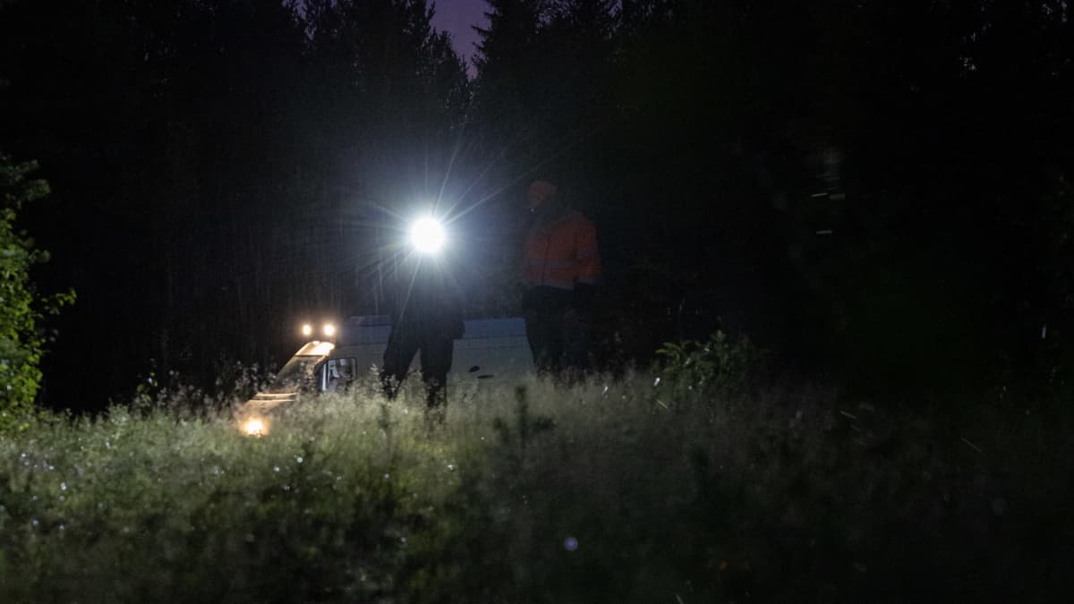Metsästäjät pimeässä maastossa otsalampun kanssa. Taustalla näkyy pakettiauto.