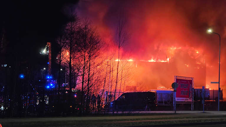 A large building in an industrial area, engulfed in flames.