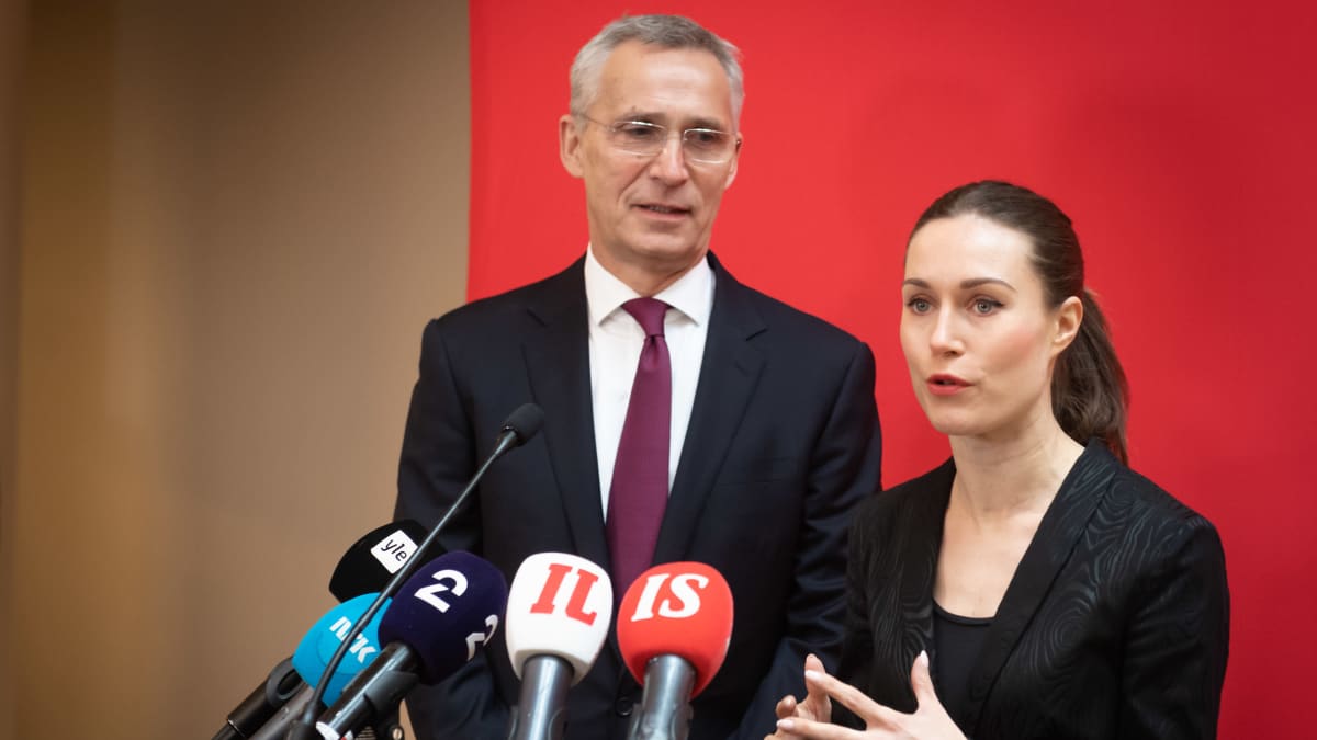 Jens Stoltenberg and Sanna Marin, both standing in dark suits behind microphones at a press conference. 