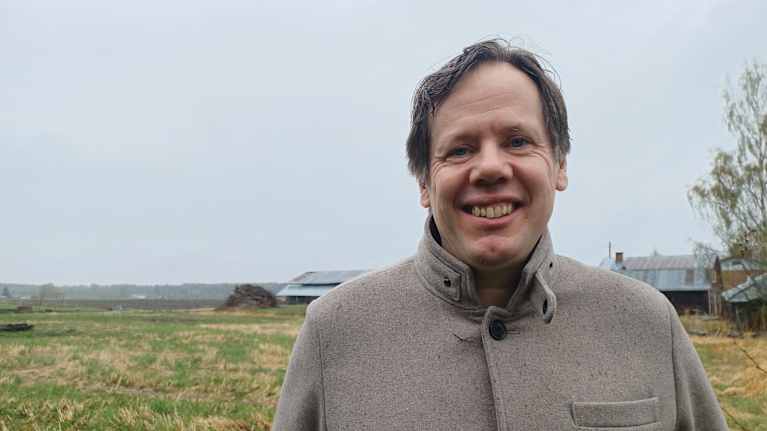 En manlig person i halvbild (fredrik Grannas) med grå vadmålskläder med ett typiskt österbottniskt landskap i bakgrunden.