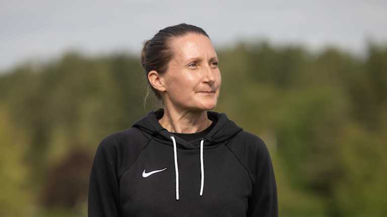 En person står utomhus på en gräsmatta med skog i bakgrunden. Personen bär en svart Nike-tröja med huva och blå jeans.