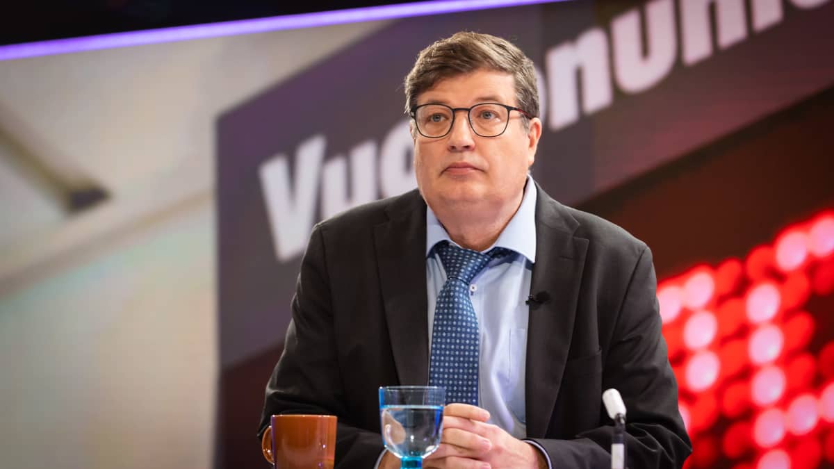A man with glasses, dark blazer and loose blue tie sits behind a table with a mug, a glass of water and a microphone. 