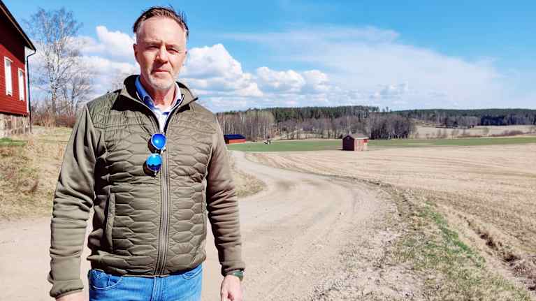 Tomas Gripenberg står vid en åker där det kör en traktor långt i bakgrunden.