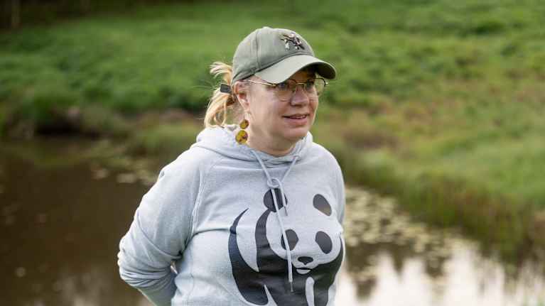 En person med en grå sweatshirt med en stor panda-logga och grå byxor. Hon står utomhus på en grön gräsmatta nära vattnet. Hon har också en keps på huvudet och gummistövlar på fötterna.