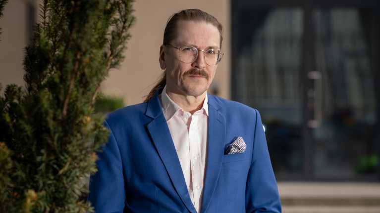 Portrait of a man with glasses, a mustache and dark hair combed back in a ponytail, wearing a blue blazer.