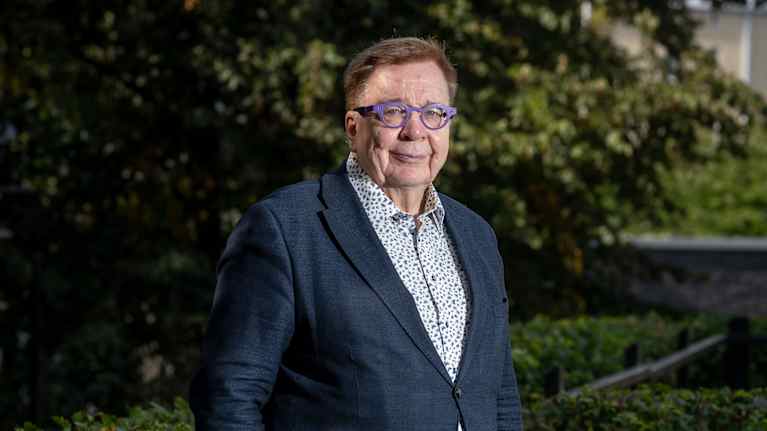 Markku Ollikainen i en mörkblå kavaj och blommig skjorta tittar in i kameran. Han står utomhus bland växter.