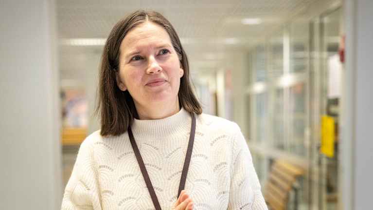 En kvinna klädd i jeans och en vit stickad tröja befinner sig i en inomhusmiljö. I bakgrunden syns en korridor med glasväggar.