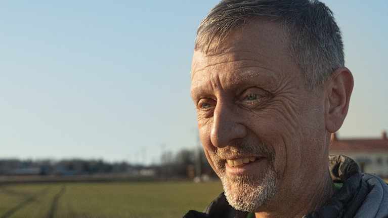 En man är klädd i en svart jacka och står på en grön åker. Det är en solig dag och ett hus syns i bakgrunden.