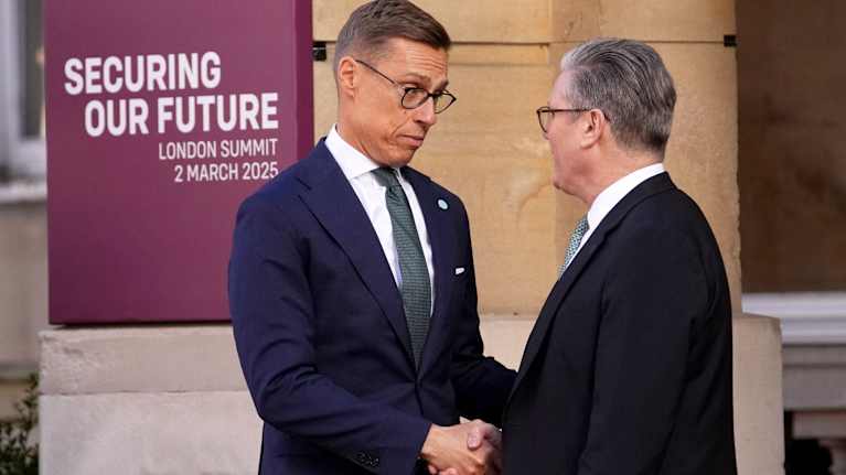 A tall man in a dark blue suit looks down into the face of a man in a black suit as they clasp hands outdoors behind a purple sign saying "SECURING OUR FUTURE LONDON SUMMIT 2 MARCH 2025".