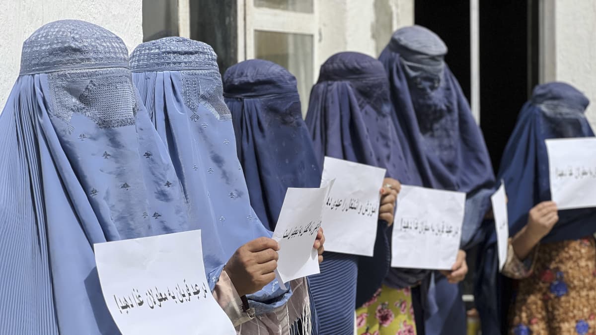 Siniseen burkaan pukeutuneet ihmiset seisovat rivissä ja pitelevät valkoisia papereita, joihin on kirjoitettu tekstiä arabialaisilla aakkosilla.