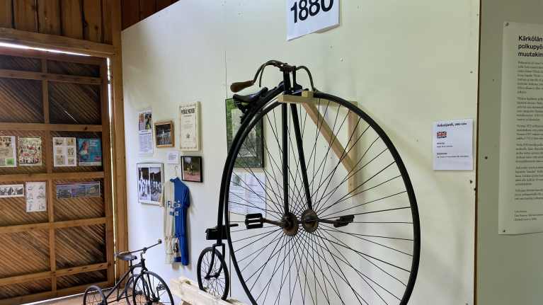 Isolla etupyörällä ja pienellä takapyörällä varustettu vanhanmallinen polkupyörä, jonka takana vastaava pienempi, lasten malli mutta kahdella pienellä takapyörällä. Seinll kehystettyjä kuvia, sinivalkoinen pyöräilypaika ja vuosiluku 18880.