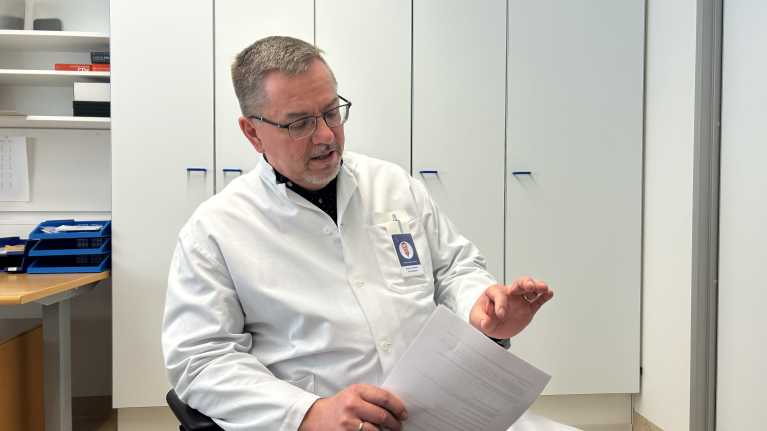 Valkoisiin pukeutunut Satasairaalan patologian osaston ylilääkäri istuu työhuoneessaan vaaleiden kaappien edessä kädessään paperinippu.