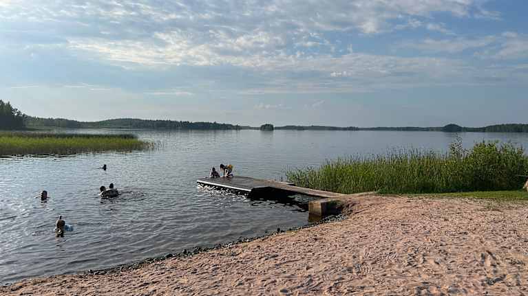 Summertime lake beach scene, with a small dock and several people swimming in the water.