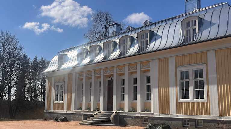 En gul och vit byggnad med mansardtak och flera fönster syns i solljus mot en blå himmel.