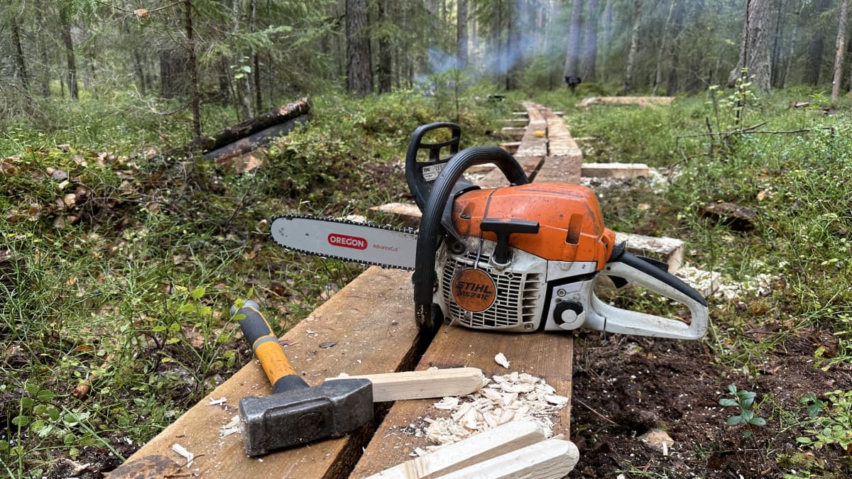 Moska, kolme puutappia ja moottorisaha sekä tuoreita lastuja asetettuna pitkospuiden päälle, taustalla näkyy nuotion savua ja metsää.