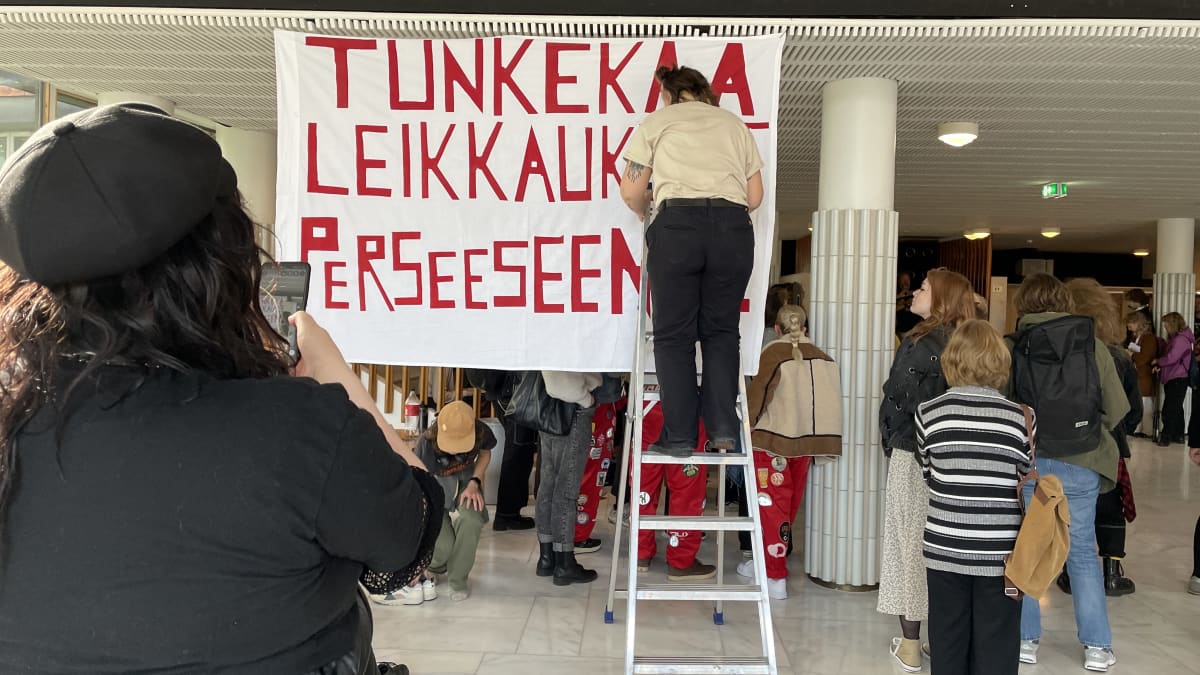 Opiskelijat kiinnittävät "Tunkekaa leikkaukset perseeseen" -lakanaa yliopiston päärakennuksen aulaan.