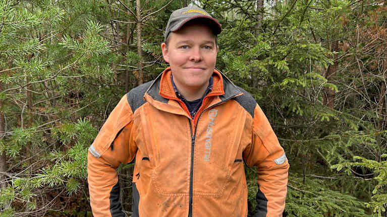 En person iförd en orange jacka står framför gröna granar i en skogsmiljö. Thomas Lindholm.