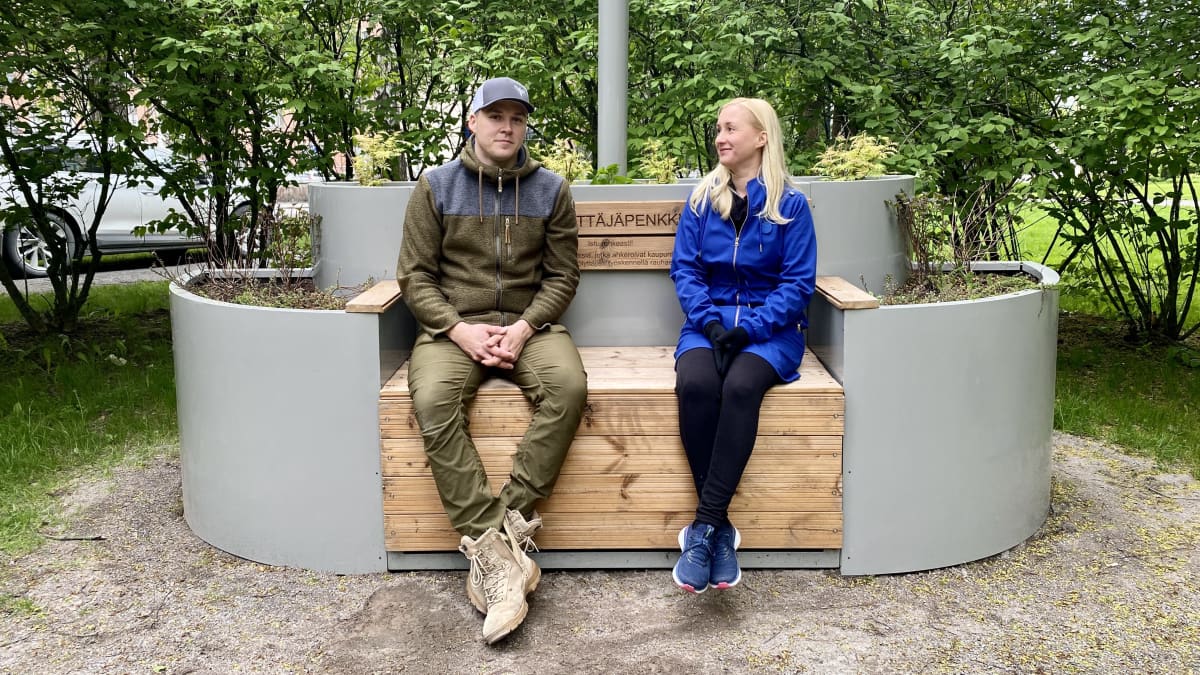 Mehiläispenkillä vasemmalla istuu maastonvihreisiin vaatteisiin pukeutunut mehiläistarhaaja Teemu Aittamaa, ja oikealla vaaleahiuksinen, sinitakkinen Liisa Jäätvuori. 