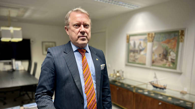 Turun yliopiston rehtori Jukka Kola nojailee työpöytäänsä tummassa liituraidallisessa puvussa ja oranssissa solmiossa.