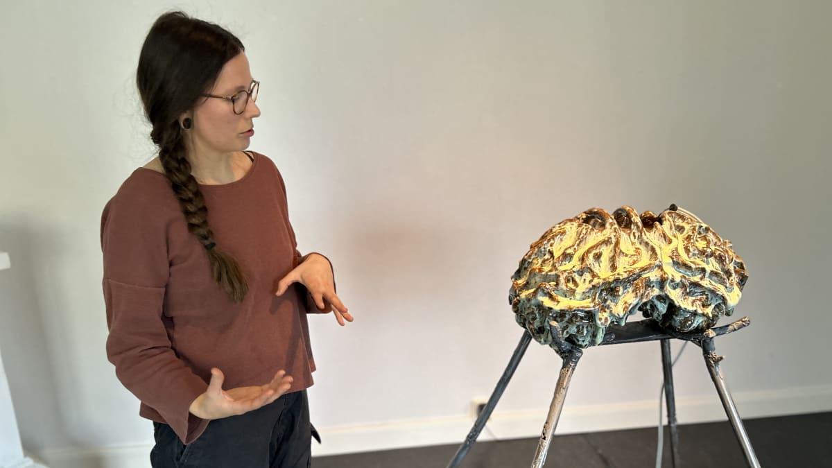 Nuori nainen esittelee keraamista taideteosta, joka seisoo neljällä jalalla taidemuseon salissa.