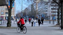 A woman with a red jacket and black helmet on a bicycle, and about 10 pedestrians in central Lahti in April, all seen from behind.