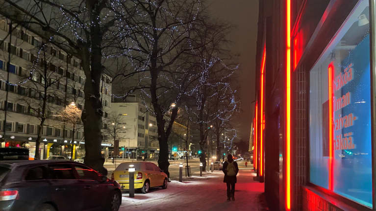 Lähikuva talvisesta kadusta kaupungin keskustassa. Kauppakeskuksen iltavalot, autoja parkissa ja liikennevalot. Ihminen kävelee poispäin.