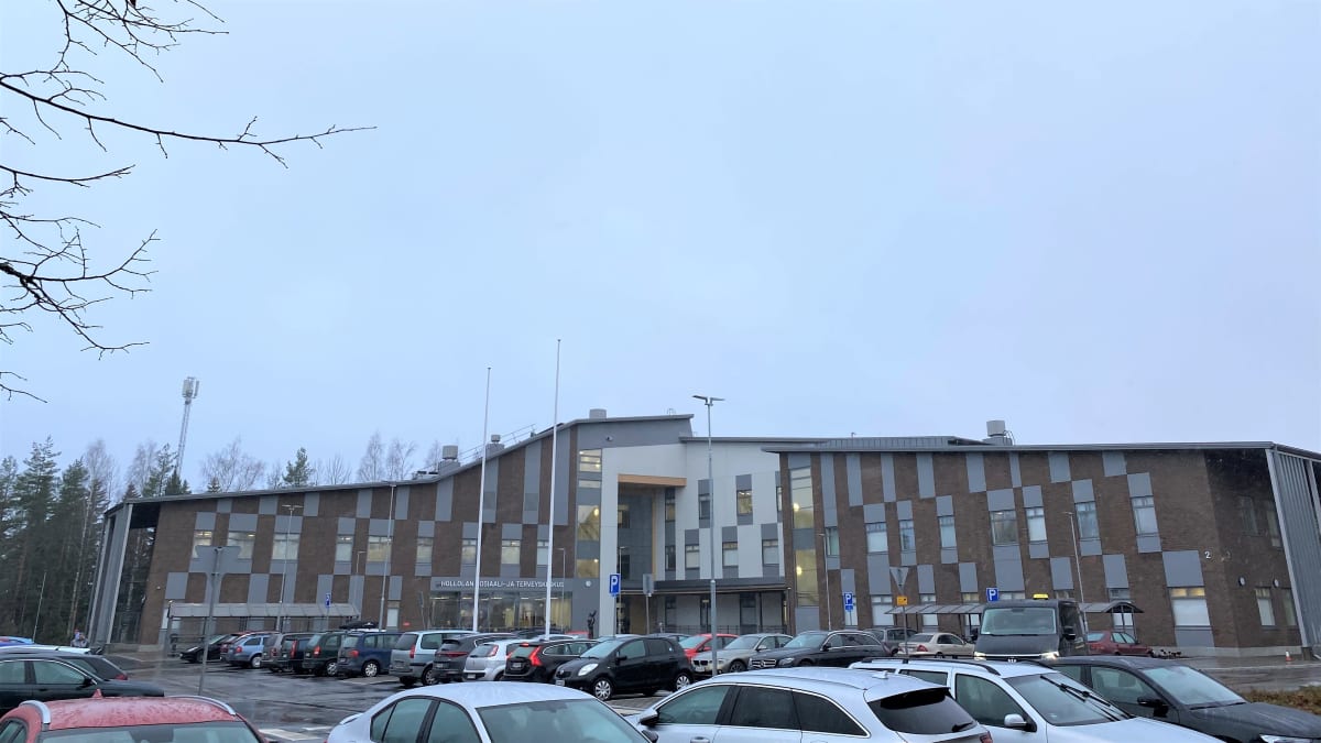 Hollolan sote-keskuksen rakennus lähikuvassa. Tummatiilinen leveä rakennus. Etualalla parkkipaikka täynnä autoja. 