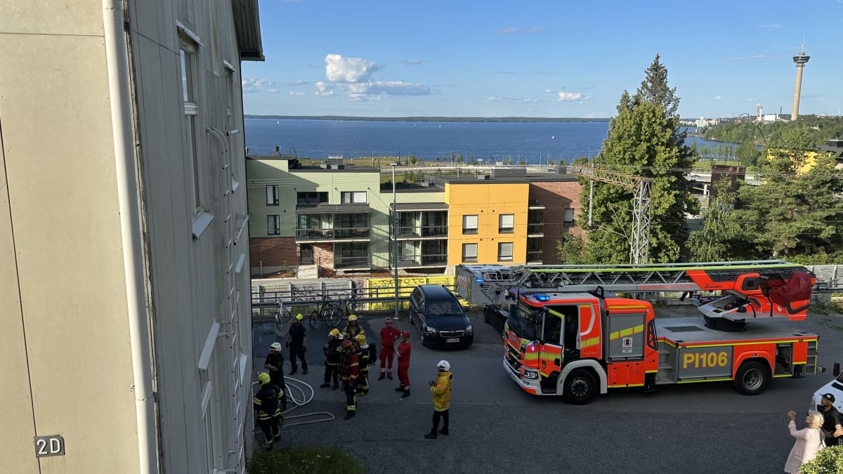 Vaalean kerrostalon edessä seisoo pelastushenkilökuntaa kantaen paloletkuja. Parkkipaikalla on yksi paloauto.