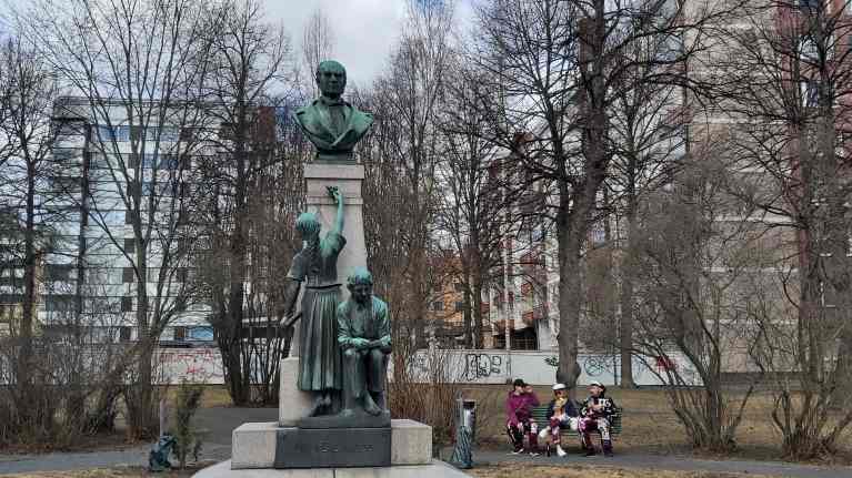 Uno Cygnaeuksen patsas Cygnaeuspuistossa Jyväskylässä, penkillä patsaan takana istuu opiskelijoita ylioppiaslakit päässä ja opiskelijahaalarit päällä.
