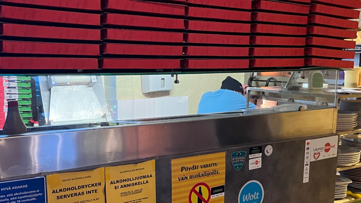 Pizzerian tiskin takana kokki paistaa pitsaa ja kuvan etuosassa on kasa pizzalaatikoita. 